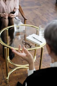 Two adults engaged in a therapy session, seated at a glass table with notebooks and water glasses.